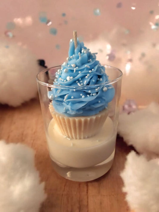 Cupcake candle with blue frosting and white sprinkles in a glass container on a wooden surface.