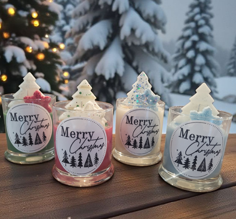 Four small Christmas tree-shaped candles with 'Merry Christmas' labels in front of a decorated Christmas tree.