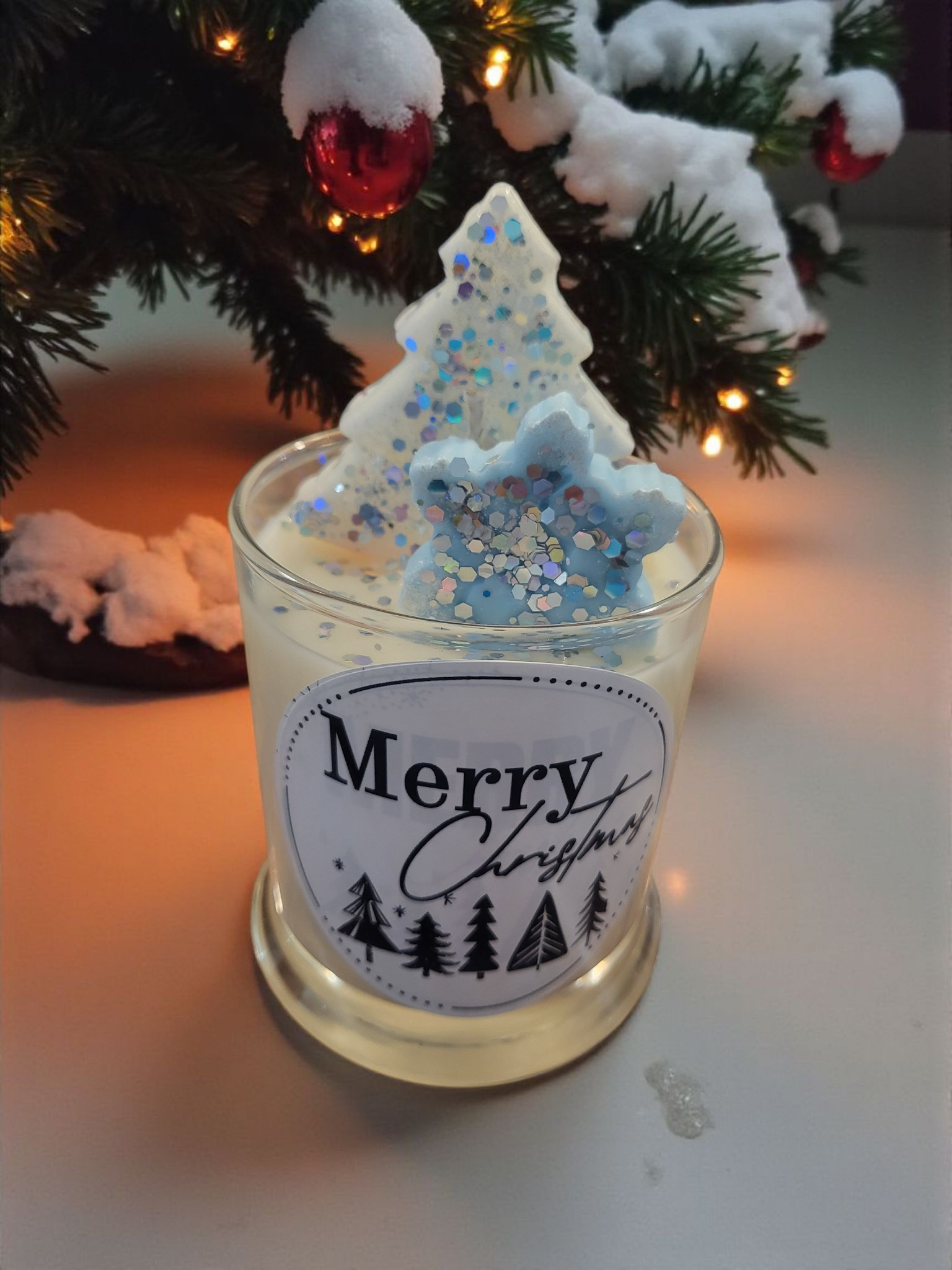 One small Christmas tree-shaped candles with 'Merry Christmas' label in front of a decorated Christmas tree.