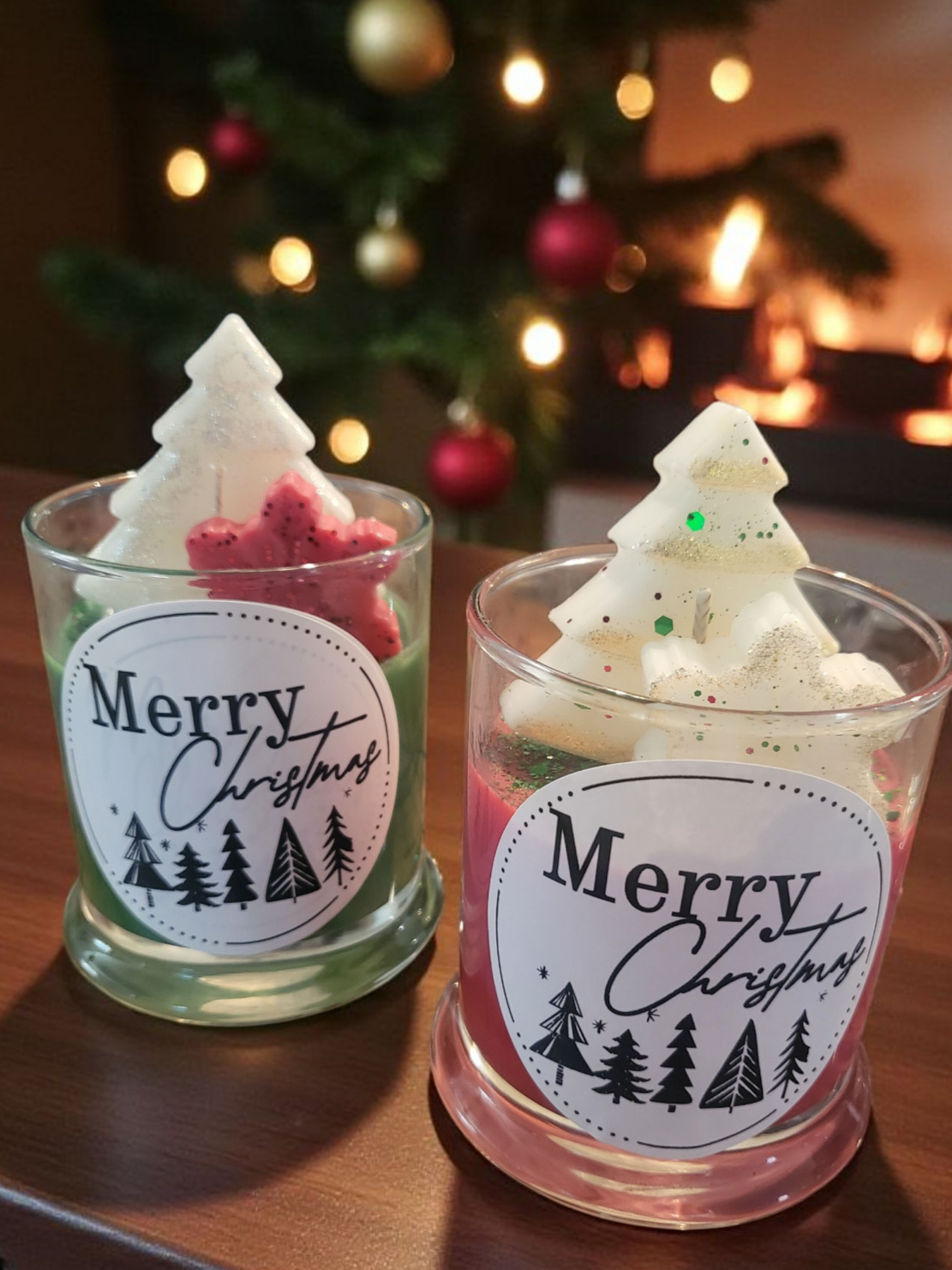 Two small Christmas tree-shaped candles with 'Merry Christmas' labels in front of a decorated Christmas tree.